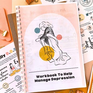 a workbook titled "Workbook To Help Manage Depression" with a colorful illustration of a person sitting on the ground. The workbook is surrounded by various papers and office supplies, including a brain illustration, a heart, and a clock.
