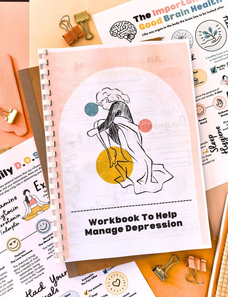 a workbook titled "Workbook To Help Manage Depression" with a colorful illustration of a person sitting on the ground. The workbook is surrounded by various papers and office supplies, including a brain illustration, a heart, and a clock.