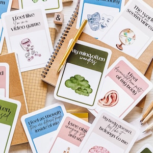 May include: A set of cards with different illustrations and text. The cards are pink, green, and blue. The text on the cards includes phrases like "I feel like I'm in a video game", "The world seems unreal around me", and "My mind often wanders".