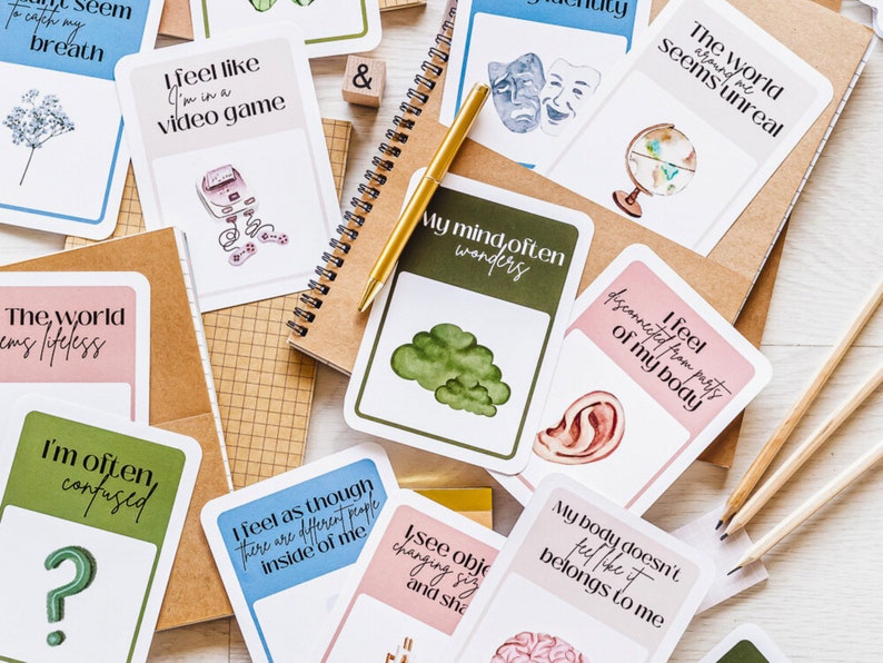 May include: A set of cards with different illustrations and text. The cards are pink, green, and blue. The text on the cards includes phrases like "I feel like I'm in a video game", "The world seems unreal around me", and "My mind often wanders".