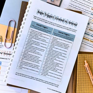 a spiral-bound notebook with a list of "anger triggers" and "internal triggers" written on it, along with some other papers and office supplies such as a stapler, paper clips, and a pen.
