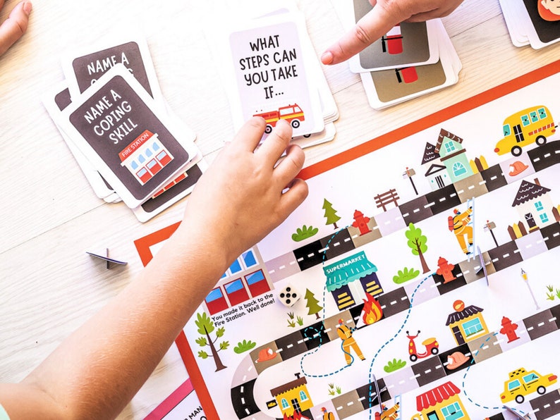 May include: A colorful board game with a fire station theme. The game board features a city street with buildings, cars, and a fire truck. The game includes cards with questions about coping skills and steps to take in different situations.