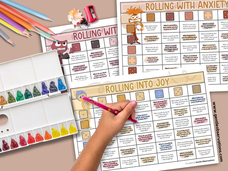 May include: A watercolor paint set, colored pencils, and three printable worksheets titled "Rolling With Anxiety" and "Rolling Into Joy" are displayed. A hand is holding a pink pencil over one of the worksheets. The worksheets have a grid format with questions.