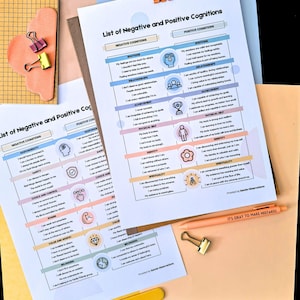 May include: Two printed sheets titled "List of Negative and Positive Cognitions" with text and illustrations, held together with orange binder clips. A peach-colored pen with the text "IT'S OKAY TO MAKE MISTAKES" is also visible. The background includes various colored papers.