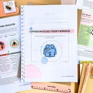a workbook titled "Compassion Focused Therapy Workbook" with a focus on practical resources for working with the Threat, Drive, and Scoring Systems. The workbook is surrounded by various papers and notes, suggesting it is part of a larger collection of materials related to compassion-focused therapy.
