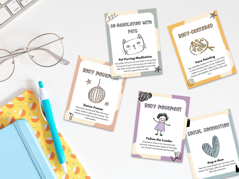 A neatly arranged stack of Somatic Therapy Coping Skill Cards for kids, featuring various emotional regulation techniques, placed on a white desk alongside a notebook and pen.
