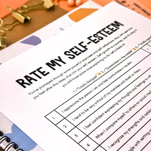 Puede incluir: Primer plano de una hoja de trabajo de evaluaci&oacute;n de la autoestima titulada "RATE MY SELF-ESTEEM". La hoja de trabajo est&aacute; en papel blanco con texto negro y una cuadr&iacute;cula para calificar las afirmaciones. Clips dorados y una regla en el fondo.