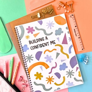 Puede incluir: Un cuaderno de espiral con el texto "BUILDING A CONFIDENT ME" y formas abstractas coloridas. La imagen incluye art&iacute;culos de papeler&iacute;a como bol&iacute;grafos, clips, una regla y chinchetas, dispuestos sobre un fondo colorido.