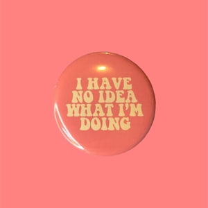 May include: A round, coral-colored button with the phrase "I HAVE NO IDEA WHAT I'M DOING" in a retro, cream-colored font. The button is centered against a solid coral background.
