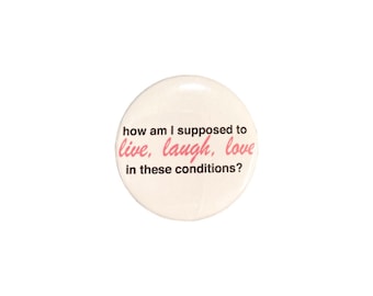 May include: A circular white pin-back button featuring black text that reads how am I supposed to and in these conditions? with the words live, laugh, love printed in a pink cursive script font centered in the middle of the button design.