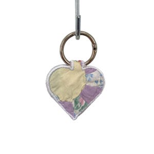 May include: A heart-shaped keychain featuring a patchwork design made of various fabric scraps in shades of pale yellow, lavender, and floral patterns. The edges are finished with white embroidery stitching, and it hangs from a silver metal ring.