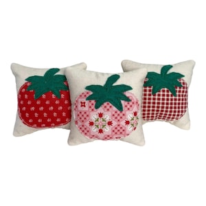 May include: Three square off-white fabric cushions, each featuring a central strawberry-shaped appliqué. The left cushion has a red floral print, the center has a pink geometric floral pattern, and the right has a red and white gingham check.