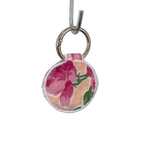 May include: A circular fabric keychain featuring a floral pattern with pink flowers, green leaves, and a peach background. The fabric is finished with white stitched edges and is attached to a silver-toned metal split ring hanging from a hook.