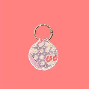 May include: A round keychain with a floral pattern in purple, red, and white. The keychain has a gold-colored metal ring for attaching keys. The background is a solid coral color.