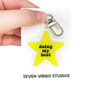 May include: A bright yellow star-shaped acrylic keychain featuring the black text "doing my best" in a rounded font. The keychain is attached to a silver-colored metal lobster clasp and is presented on a white backing card labeled "SEVEN VIRGO STUDIOS".
