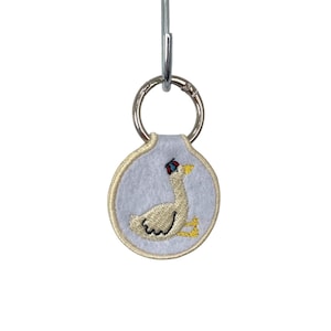 May include: A circular light blue felt keychain featuring an embroidered white goose wearing a small red and blue hat. The goose has yellow feet and a yellow beak. The keychain is attached to a silver metal split ring and a lobster clasp.