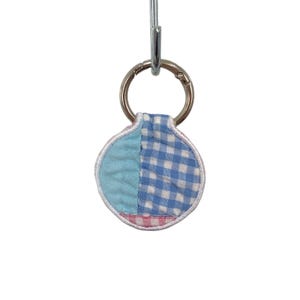 May include: A circular fabric keychain featuring a patchwork design of light blue solid fabric, blue and white gingham, and a small section of pink and white gingham at the bottom, all finished with a white stitched border and a silver metal ring.