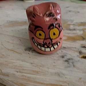 May include: A pink ceramic Cheshire Cat-themed container. The container features a smiling face with yellow eyes and a wide grin. The container is hand-painted with black outlines and details.