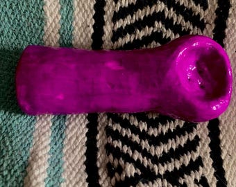 Puede incluir: Una pipa de color magenta vibrante, de forma cilíndrica, con un cuenco en un extremo. Descansa sobre un textil tejido con un patrón negro, blanco y turquesa. La superficie de la pipa es lisa y brillante.