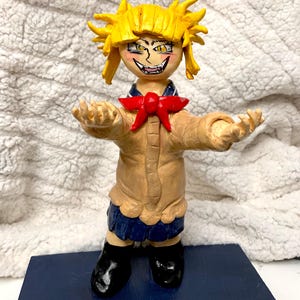 May include: A handmade figurine of a character with spiky yellow hair, a red bow, and a tan jacket over a blue skirt. The figure has a menacing expression and is standing on a blue base. Black boots complete the look.