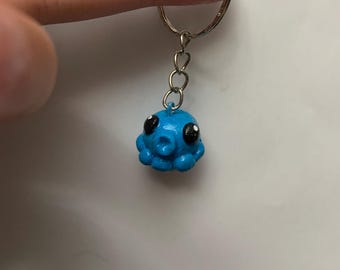 Könnte beinhalten: Ein blauer Oktopus-Schlüsselanhänger mit schwarzen Augen und einem silbernen Ring. Der Oktopus ist ein kleiner, handgefertigter Anhänger mit strukturierter Oberfläche. Der Schlüsselanhänger ist an einem silbernen Metallring befestigt.
