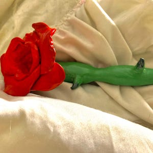 Könnte beinhalten: Eine leuchtend rote Rosenskulptur neben einer grünen Eidechsenskulptur. Die Rose steht in voller Blüte, und die Eidechse hat ein dunkelgraues Horn. Beide sind auf einem cremefarbenen Stoff platziert.
