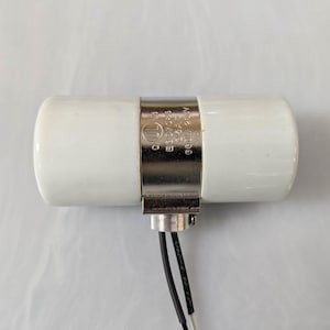 May include: A white ceramic light socket with a metal band. The band has text and a logo. Two wires extend from the base. The socket is designed to hold a light bulb.