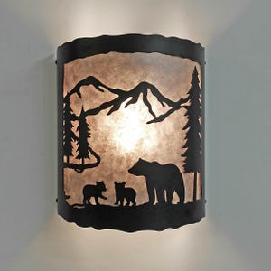 May include: A rustic wall sconce with a black metal silhouette of a bear family in a forest scene. The design includes mountains, trees, and a warm, glowing light source, creating a cozy ambiance. The sconce is mounted on a white wall.
