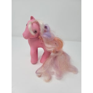 May include: A pink plastic toy pony figure with a long, flowing mane and tail featuring blended strands of white, lavender, and peach-colored hair. The pony has large, dark eyes and a small, sculpted symbol on its flank, standing in a fixed pose.