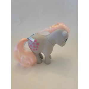 May include: A gray toy pony with a pink mane and tail. Pink heart shapes decorate the pony's side. A light blue ribbon is tied around the tail. The pony has blue eyes and is a children's toy.