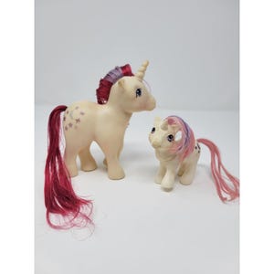 May include: Two cream-colored unicorn figurines with long, rooted hair. The larger unicorn has a dark pink and purple mane and tail with a moon and star symbol on its flank. The smaller unicorn has a pink and purple mane and tail with stars.
