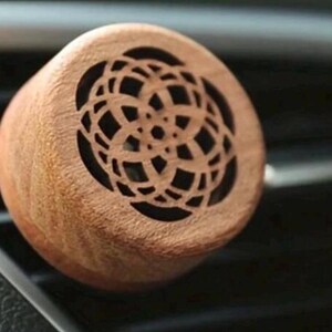 May include: A circular wooden car air freshener with an intricate, open pattern. The light brown air freshener is positioned in a car's air vent. The background is dark, highlighting the product's design.