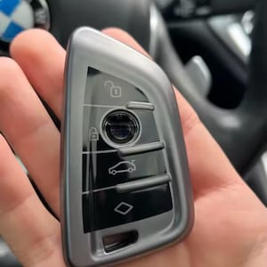 May include: A gray car key with black accents, featuring buttons for lock, unlock, and other functions. The key has a sleek, modern design and is held in a hand.