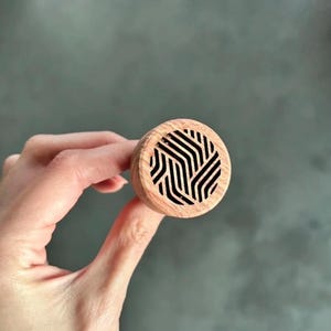 Może przedstawiać: Drewniany korek do butelki o okrągłym kształcie. Korek ma jasnobrązowe usłojenie drewna i czarny geometryczny wzór na górze. Korek jest trzymany w dłoni na szarym tle.