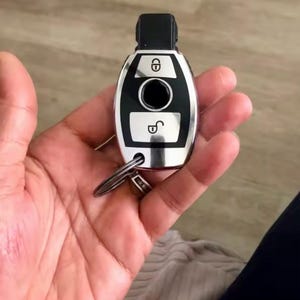 Op de afbeelding: Een zilveren en zwarte autosleutel met een sleutelring. De sleutel heeft een zwarte bovenkant en een zilveren behuizing met vergrendel- en ontgrendelsymbolen. De sleutel wordt in een hand gehouden.