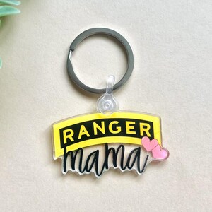 May include: A clear acrylic keychain with a silver keyring. The keychain has a yellow banner with the word "RANGER" in black and the word "mama" in a script font. Two pink hearts are on the right.