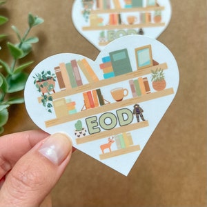 May include: White heart-shaped sticker with a bookshelf design. The shelves are filled with books, plants, and small decorative items. The text "EOD" is written in green letters on the sticker.