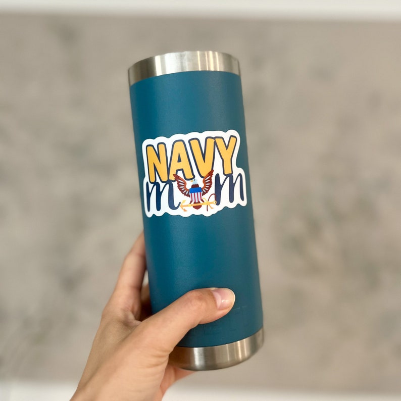 May include: A teal blue tumbler with a white sticker that says "NAVY MOM" in yellow letters with a small American flag graphic in the center.