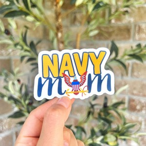 May include: White sticker with the text "NAVY mom" in blue and yellow lettering. A small image of an eagle with an American flag is in the center of the sticker.