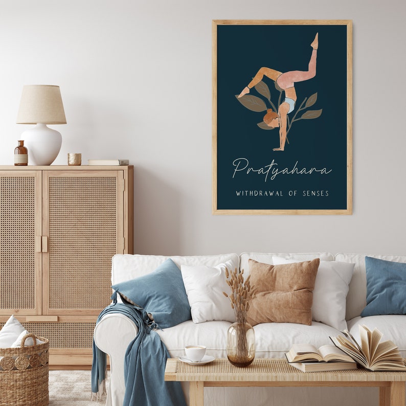 May include: A framed art print featuring a yoga pose illustration against a dark blue background. The artwork includes the word "Pratyahara" and the phrase "Withdrawal of Senses" in elegant script. The print is displayed in a living room setting.