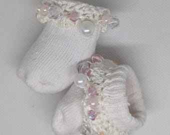 May include: A pair of white baby socks with crocheted cuffs. The cuffs are decorated with pink and clear beads, and large white pearls. The socks have a delicate, feminine design.