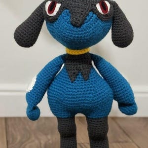 Könnte beinhalten: Handgemachte blaue und graue Häkelpuppe mit roten Augen und gelbem Kragen. Die Puppe hat ein markantes Design mit maskenartigen Merkmalen und spitzen Ohren. Die Puppe steht auf einem Holzboden.