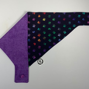 May include: A reversible dog bandana with a black background and rainbow paw prints on one side and a solid purple fabric on the other side. The bandana has a snap closure.