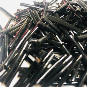 Pode incluir: Um monte de contas de vidro preto, cada conta é um cilindro longo com um furo no centro. As contas são perfeitas para fazer joias ou outros projetos de artesanato.