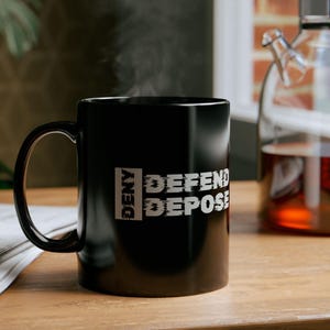 May include: Black coffee mug with white text that reads "Deny Depose Defend". The mug is sitting on a wooden table with a glass teapot in the background.