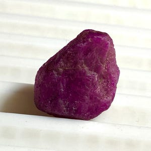 May include: A rough, uncut ruby gemstone. The ruby is a deep, vibrant magenta color with a slightly textured surface. The gemstone is displayed against a white background.