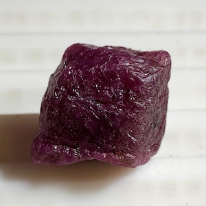 May include: A rough, uncut ruby gemstone. The ruby is a deep, rich red color with a slightly uneven surface. The gemstone is a small, irregularly shaped rock.