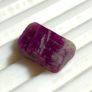 Peut inclure: Une pierre précieuse de rubis brute et non taillée, de couleur magenta foncé. Le rubis a une surface rugueuse et texturée et une forme rectangulaire. La pierre est présentée sur une surface blanche et striée, mettant en valeur sa beauté naturelle.