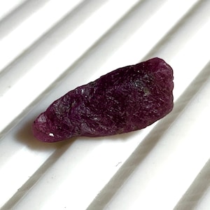 May include: A raw ruby gemstone in a deep burgundy hue. The uncut stone has a rough, textured surface and is set against a white, ridged background. The gemstone is a natural, unpolished specimen.