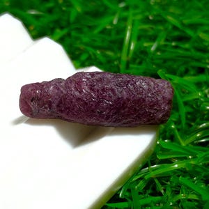 Puede incluir: Una gema de rubí en bruto, de color burdeos intenso, descansa sobre una superficie blanca, sobre un fondo de césped artificial verde vibrante. La textura rugosa y el color intenso del rubí se destacan.